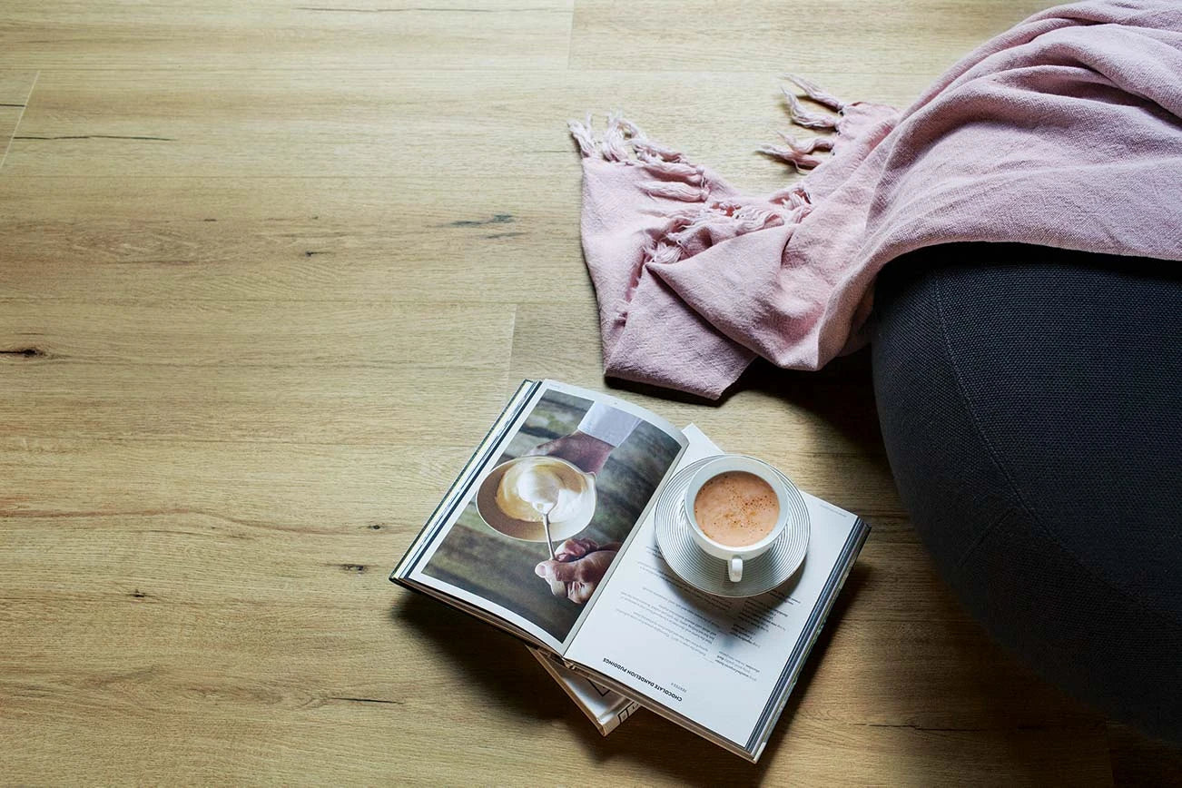 Natural Oak Vinyl Plank Sahara Dune underfoot with an open recipe book, a coffee cup and a pink throw